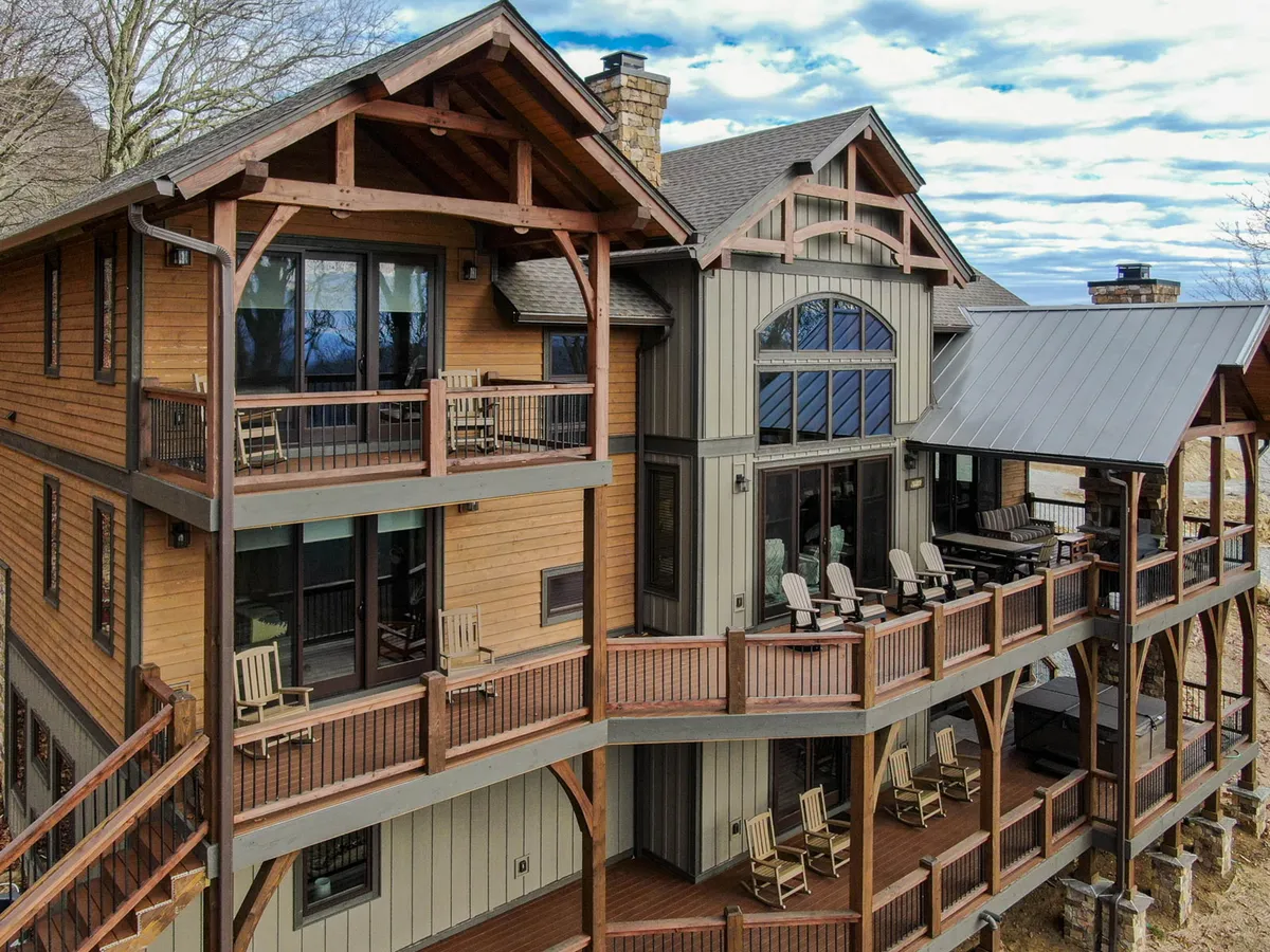 The hero image showcases the impressive exterior of 5 Cubs Lodge, with expansive wooden balconies perfect for gathering, large windows that reveal glimpses of the inviting interiors, a striking stone chimney, and comfortable outdoor furniture. The scenic mountain backdrop sets a tranquil tone, while the golden sunset bathes the home in a warm, welcoming glow, hinting at the perfect spot for evening relaxation or family celebrations.