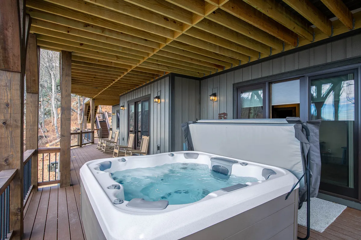 An inviting outdoor hot tub sits on a wooden deck, surrounded by comfortable seating and embraced by tranquil mountain scenery. The setting is serene, perfect for unwinding after a day of adventure and soaking in the views of the Blue Ridge peaks.