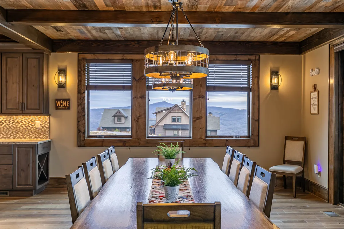 The image centers on an expansive wooden dining table set beneath elegant chandeliers, with windows offering scenic views. The table is large enough for group celebrations, intimate family dinners, or lively game nights.