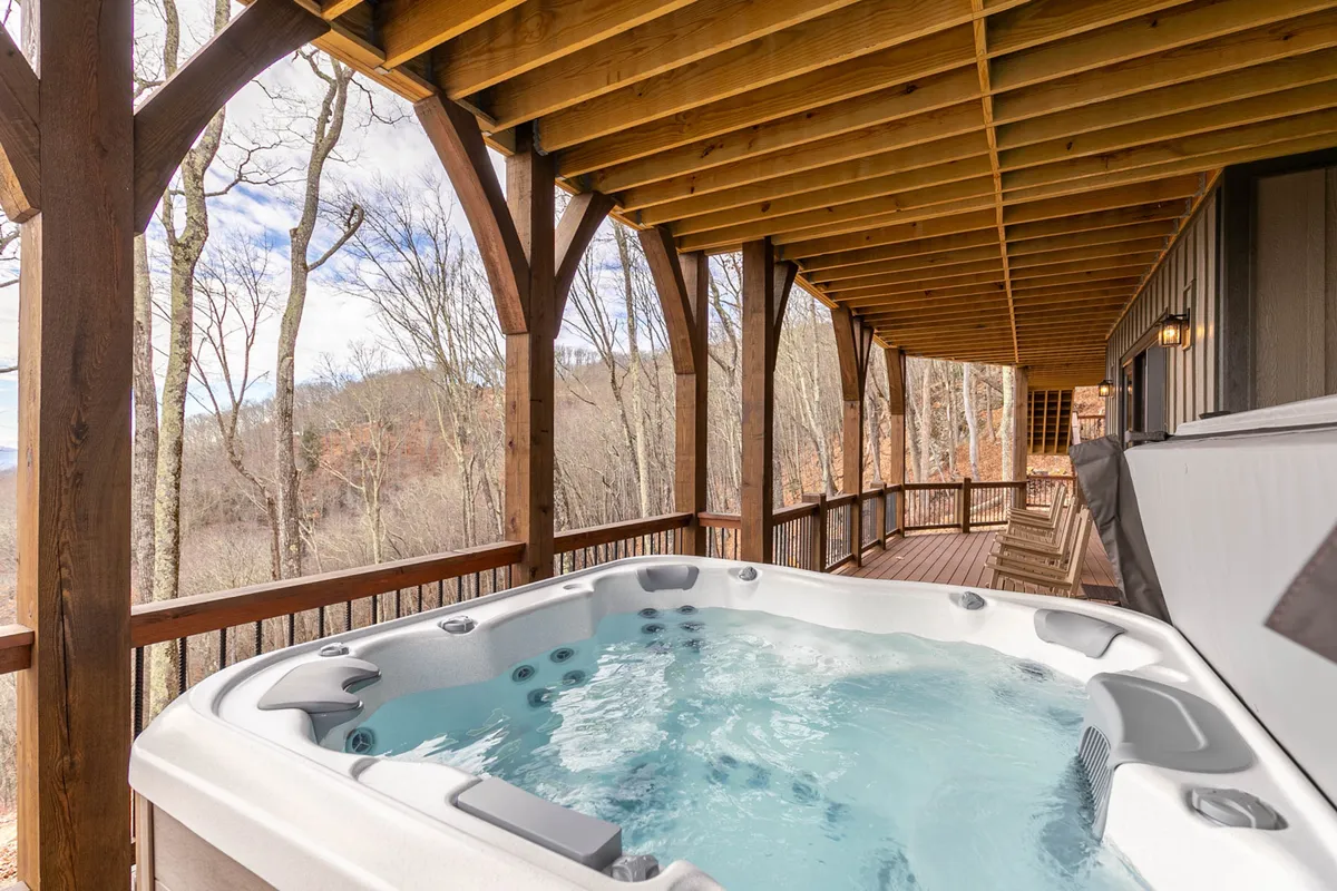 An exterior deck view with a bubbling hot tub framed by wooden beams and surrounded by nature. The tranquil scene invites relaxation, with space for friends or family to unwind while enjoying the outdoors, day or night.