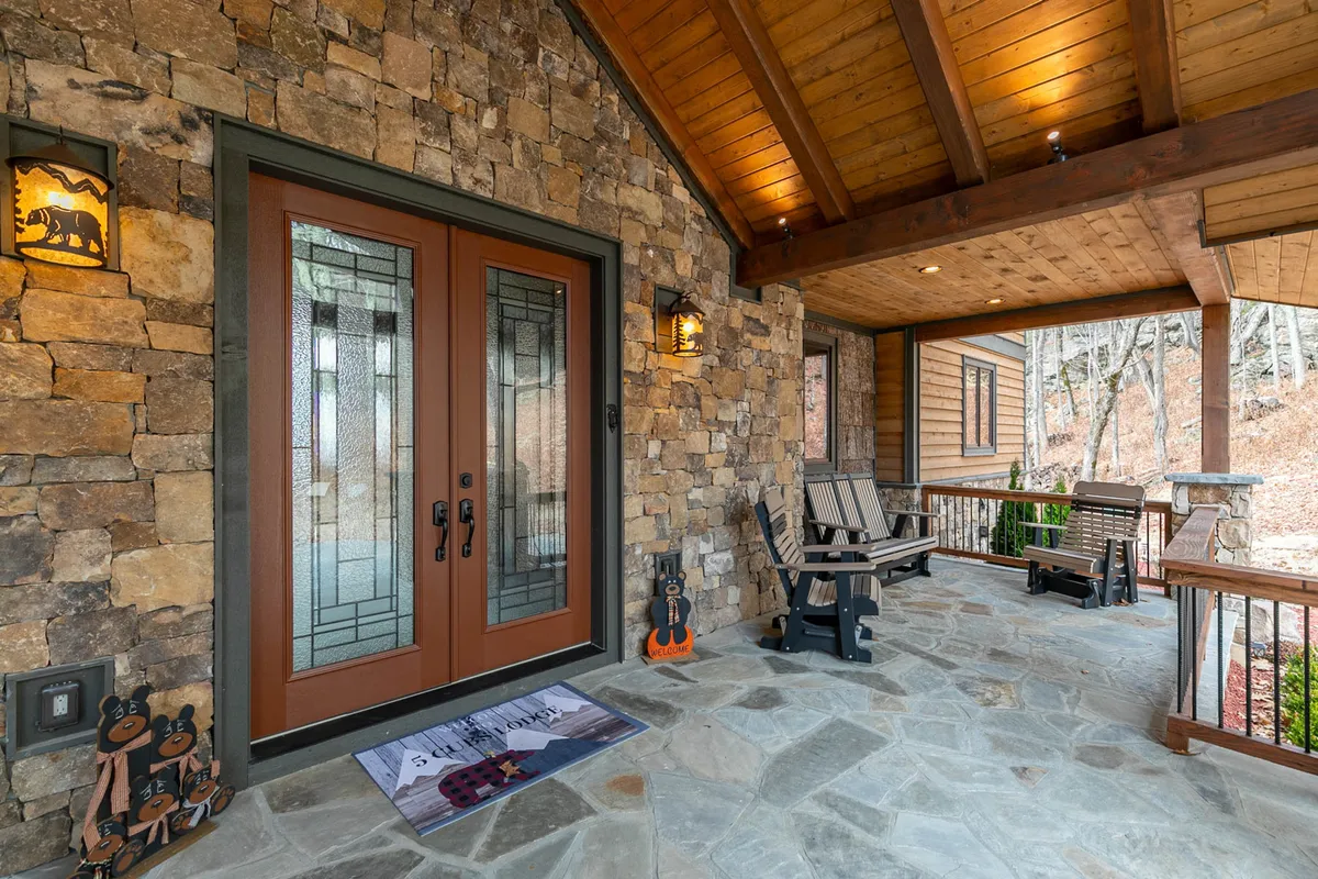 This is an exterior image showcasing the welcoming entryway to 5 Cubs Lodge, featuring double doors with stained glass, a stone wall, a wooden ceiling, cozy rocking chairs, and decorative bear figures. The porch area looks inviting, surrounded by natural scenery, and suggests a peaceful retreat setting.
