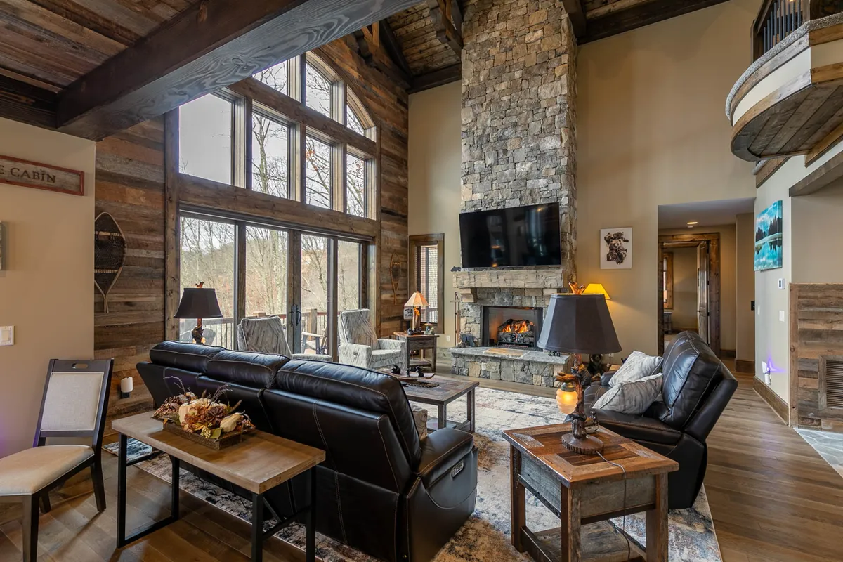 This image showcases the main living room, featuring a majestic stone fireplace, soaring wooden beams, large windows that invite natural light, and plush leather sofas arranged for conversation or relaxation. The overall setting is inviting and luxurious, capturing the essence of grand gathering spaces.