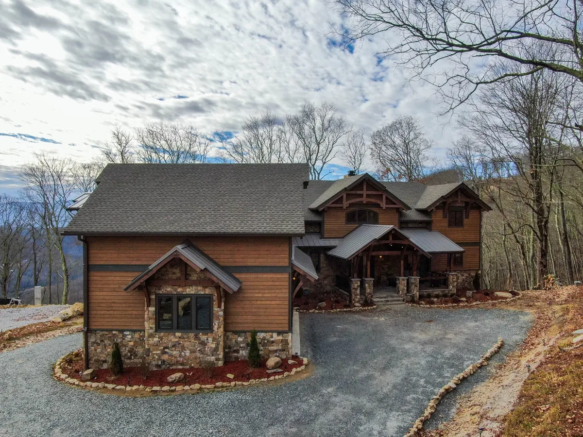 A welcoming exterior view of the lodge, showcasing its unique architectural design, stonework, and expansive driveway. The house is set amidst natural surroundings, evoking a sense of space and opportunity for gatherings or outdoor events.