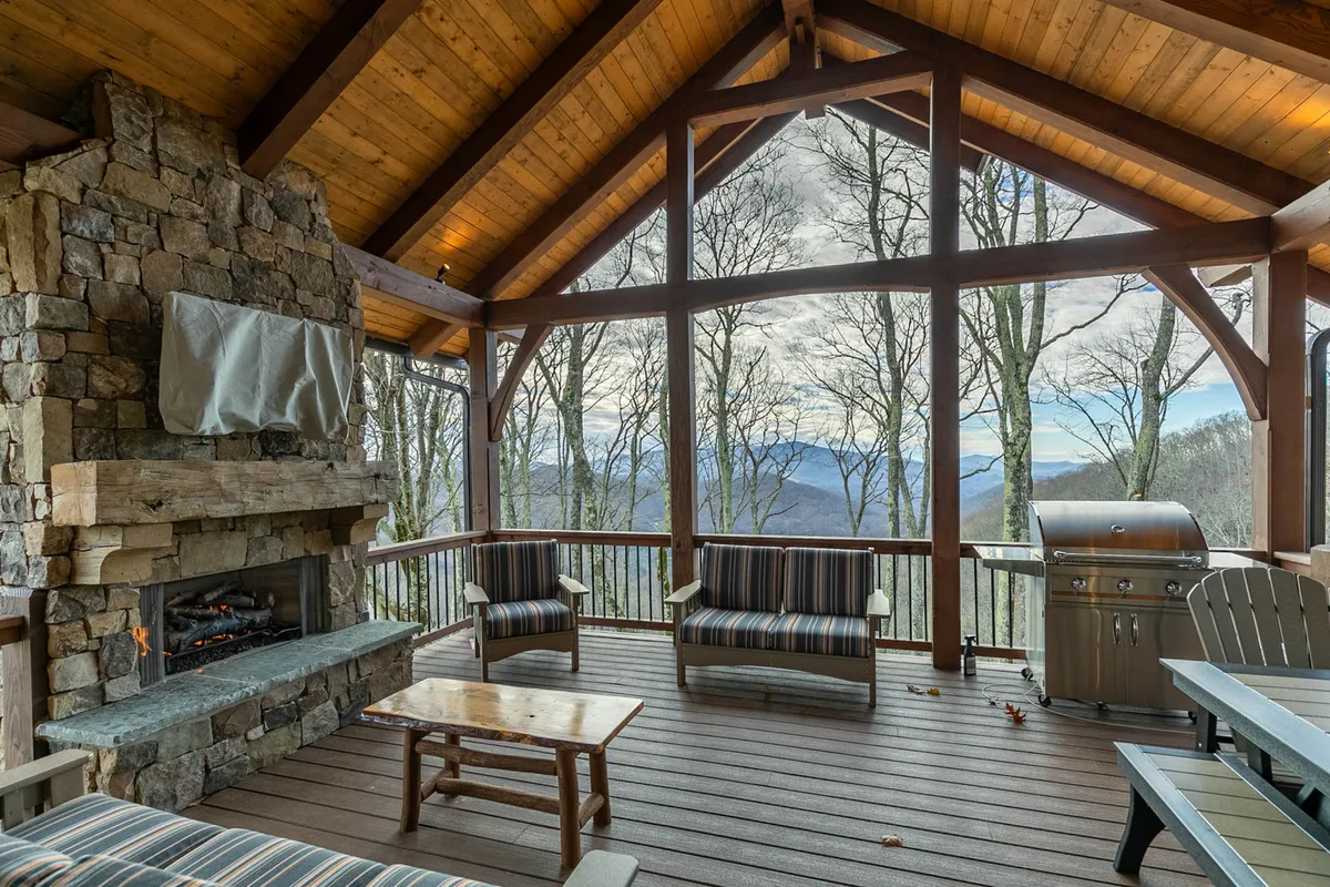 A spacious wooden deck with comfortable seating and a stone fireplace. The deck offers both covered and open-air areas, large windows framing panoramic mountain views, and a grill area for outdoor meals.